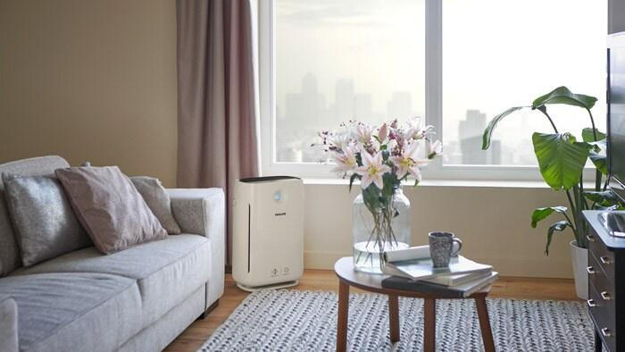 Modern living room with an air purifier positioned near a sofa and large window, promoting cleaner indoor air and improved home air quality.